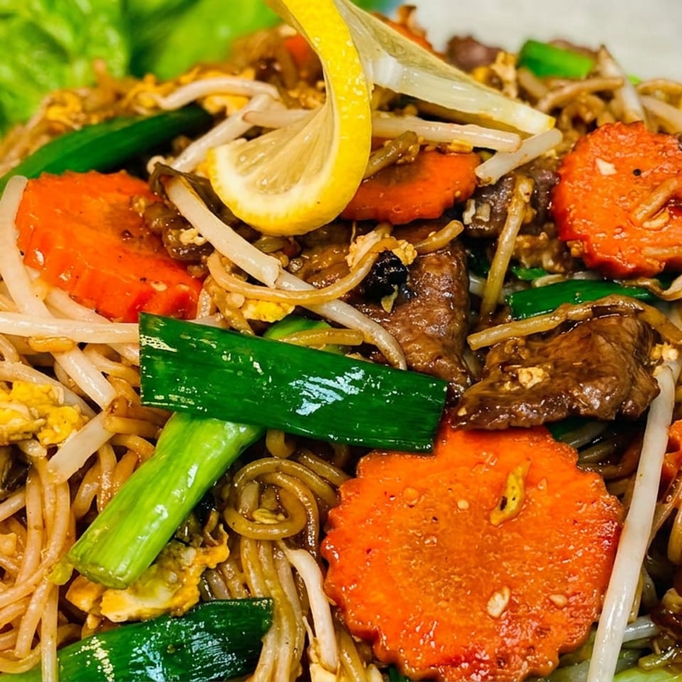 Stir-fried noodles with beef, carrots, bean sprouts, scallions, scrambled egg, and lemon garnish on a plate.