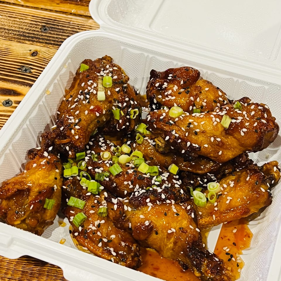 Glazed chicken wings topped with sesame seeds and scallions in a takeout box.