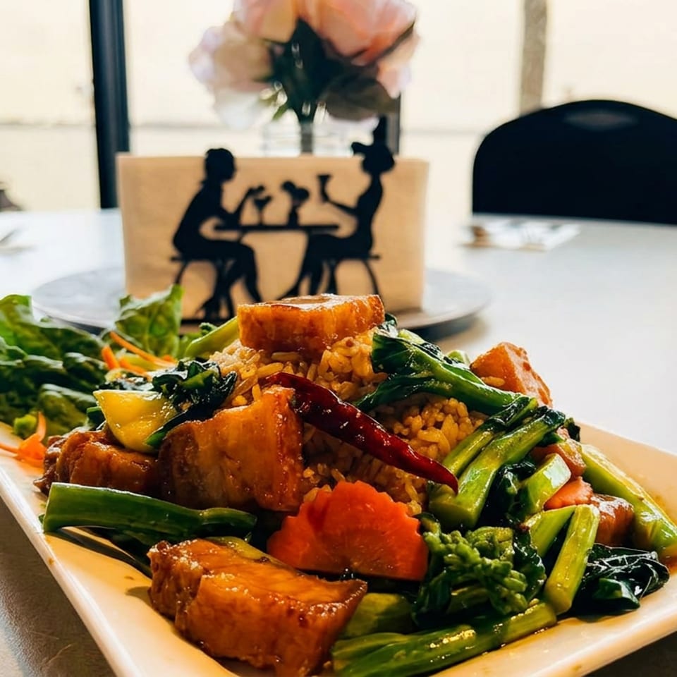 Colorful stir-fried rice with vegetables and glazed tofu or meat served on a rectangular plate in a bright restaurant.
